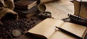 Historisch arrangiertes Stillleben mit Kaffeebohnen, einem Maria-Theresien-Taler, Kompass, Kartenmaterial, Büchern, einem Modellsegelschiff sowie einem offenen Notizbuch mit Füllfeder auf einem Holztisch