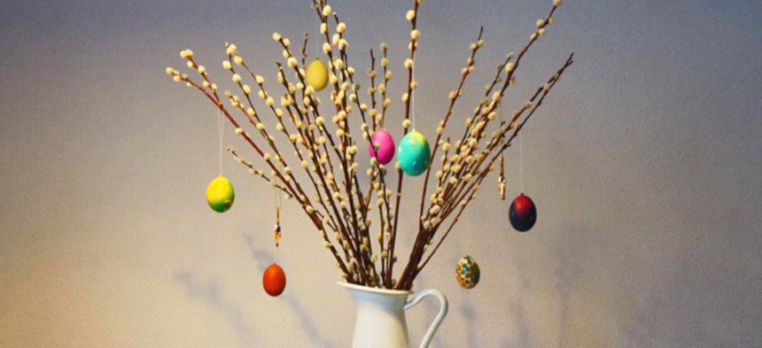 In einem weißen Krug steht ein Weidekätzchenstrauß, an dem bunte Ostereier hängen.