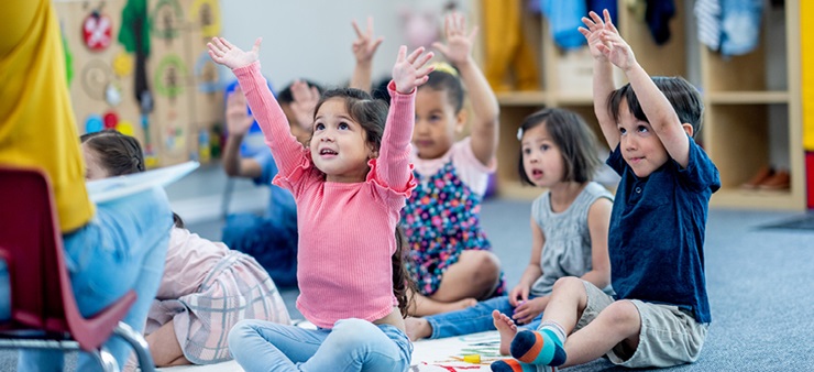 Kinder sitzen auf dem Boden und schauen gespannt hoch Richtung Erzieherin. Sie ziehen die Arme in die Höhe. 