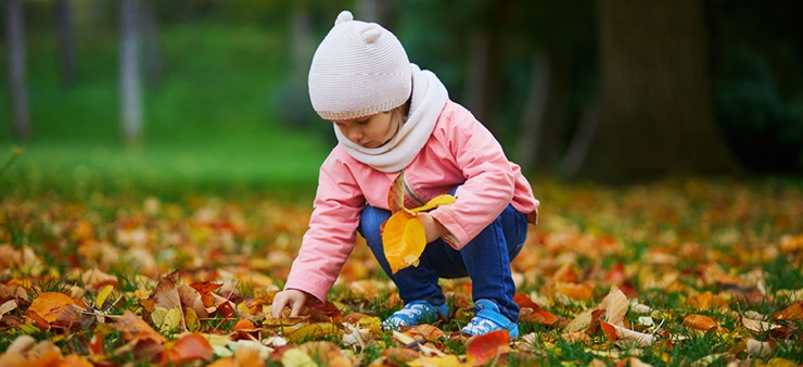 Von buntem Herbstlaub umgeben sehen wir wie ein Kind in dicker Jacke und mit Mütze die bunten Blätter aufhebt.