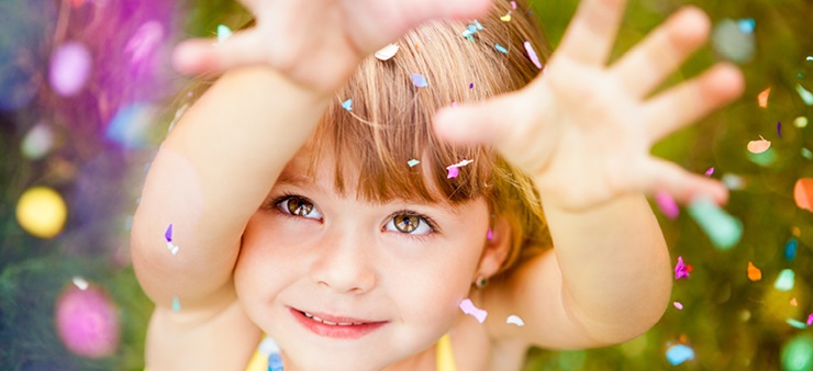 Das Bild zeigt ein Kind aus Vogelperspektive. Es schaut lächelnd hinauf und versucht bunte Konfetti zu fangen, das um es herum zu Boden sinkt.