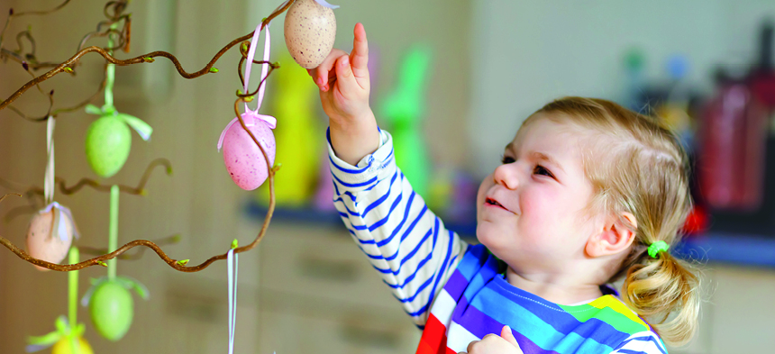 An mehreren Ästen hängen bunte Ostereier. Rechts davon steht ein kleines Mädchen, das fasziniert die Ostereier begutachtet und anfasst.