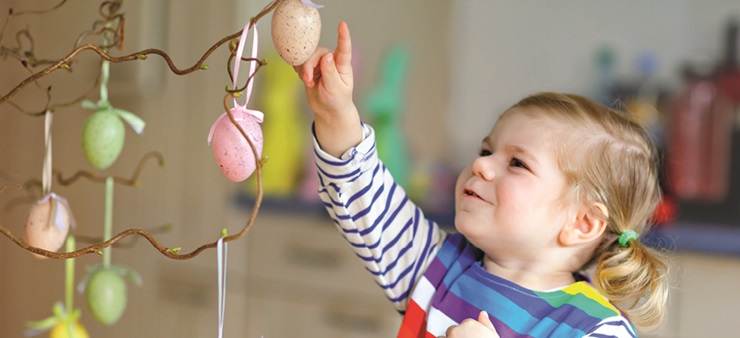 An mehreren Ästen hängen bunte Ostereier. Rechts davon steht ein kleines Mädchen, das fasziniert die Ostereier begutachtet und anfasst.
