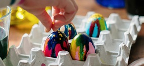 Bunte Ostereier stecken in einer Schachtel, eine Kinderhand greift nach dem linken Ei.