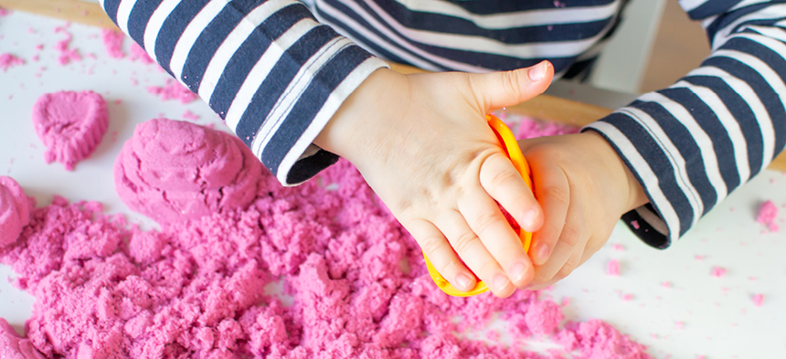 Aus der Vogelperspektive sehen wir Kinderhände, die mit pinker Knete spielen. Die Finger drücken die Knete in eine Form. Auf dem Tisch ist die Knete zu sehen.