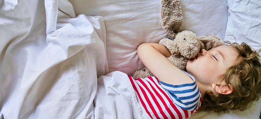 Längs liegend ist ein Junge in einem rot-blau gestreiften T-Shirt zugedeckt im Bett zu sehen. Er umarmt einen Plüsch-Hasen und schläft. 
