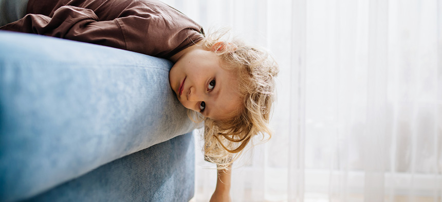 Ein Kind liegt auf dem Bauch auf einem blauen Sofa, lässt die Hände und den Kopf herunterbaumeln und schaut frustriert.