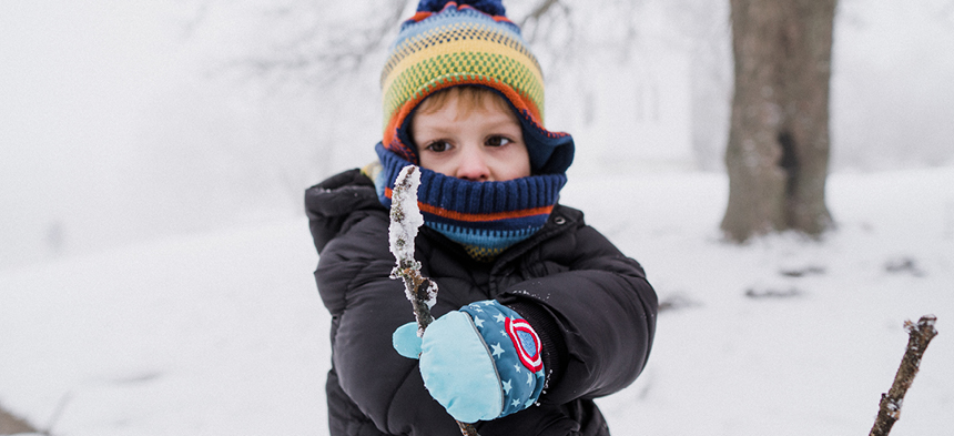 Wir sehen einen blonden Jungen in der Mitte des Bildes. Er trägt eine bunte Mütze, blaue Handschuhe, einen blau-roten Schal und eine dicke, schwarze Winterjacke. Der Junge hält einen vereisten Stock in der Hand und schaut ihn konzentriert an. Im Hintergrund eine Schneelandschaft.
