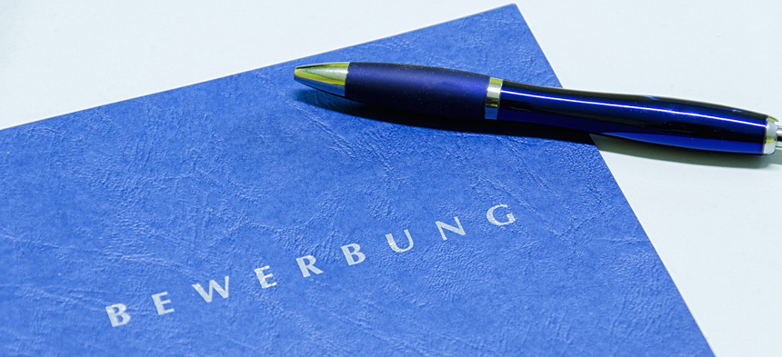 Eine blaue Mappe mit der Aufschrift "Bewerbung" liegt auf einem weißen Tisch, darüber ein blauer Kugelschreiber.
