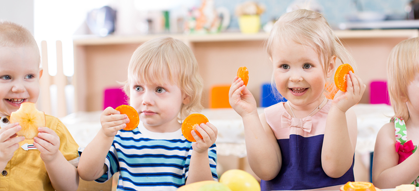 3 Kinder sitzen am Tisch und halten freudig Orangenscheiben in der Hand. 
