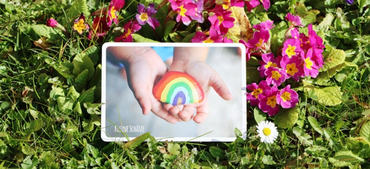 Auf einer Blumenwiese liegt eine Karte. Die Abbildung auf der Karte zeigen Kinderhände, die einen Stein halten, auf dem ein Regenbogen gemalt ist.