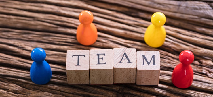 Vier Holzwürfel, auf denen Buchstaben abgedruckt sind, bilden das Wort "Team", daneben stehen vier bunte Figuren.