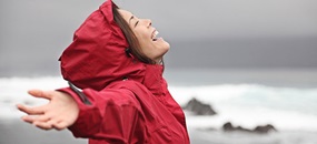 Eine junge Frau steht am Meer und lacht glücklich gen Himmel.