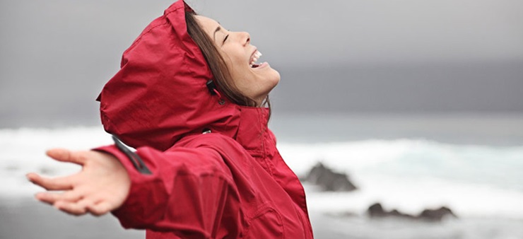 Eine junge Frau steht am Meer und lacht glücklich gen Himmel.