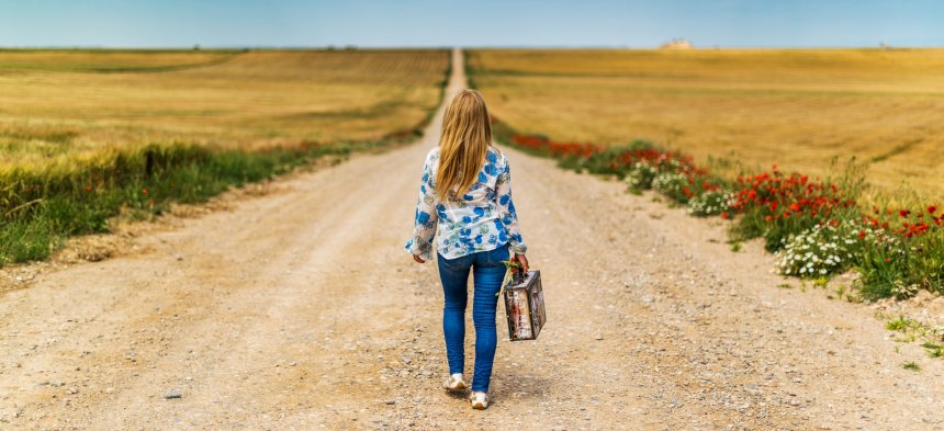 Eine Frau mit Koffer geht einen langen Pfad Richtung Horizont entlang.