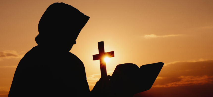 Die schwarze Silhouette eines Mönchs mit Kapuze hält ein aufgeschlagenes Buch und ein Kruzifix in der Hand, im Hintergrund ein Sonnenuntergang.