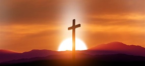 Ein Kreuz steht vor einer Berglandschaft, hinter der die Sonne untergeht.