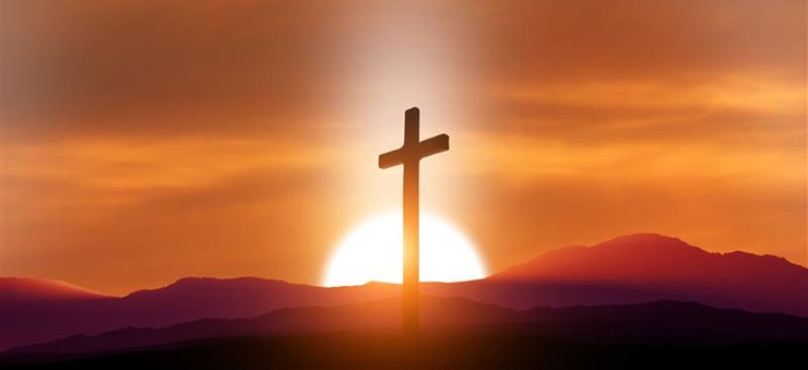 Ein Kreuz steht vor einer Berglandschaft, hinter der die Sonne untergeht.
