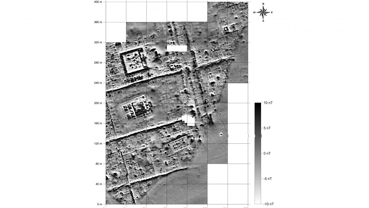 Graustufenaufnahme einer archäologischen Ausgrabungsfläche mit einem Raster und Maßstabsbalken. Mehrere rechteckige und quadratische Strukturen sind erkennbar, die wie Grundrisse von Gebäuden wirken. Deutliche Linien verlaufen diagonal und horizontal durch das Bild und deuten auf Straßen oder Wege hin. In der rechten oberen Ecke befindet sich eine Windrose zur Orientierung. Die Bildfläche zeigt unterschiedliche Helligkeitswerte, die vermutlich verschiedene Materialien oder Bodenschichten markieren.
