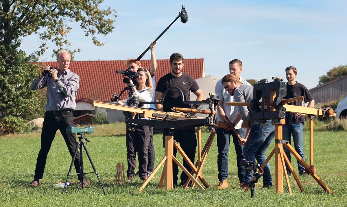Foto von sieben Personen in Freigelände, die hinter dem Nachbau zweier Geschütze stehen; ein Mann mit Fernglas, eine Frau mit Kamera, ein Mann mit Mikrofon. Im Hintergrund ist ein Hausdach zu sehen.