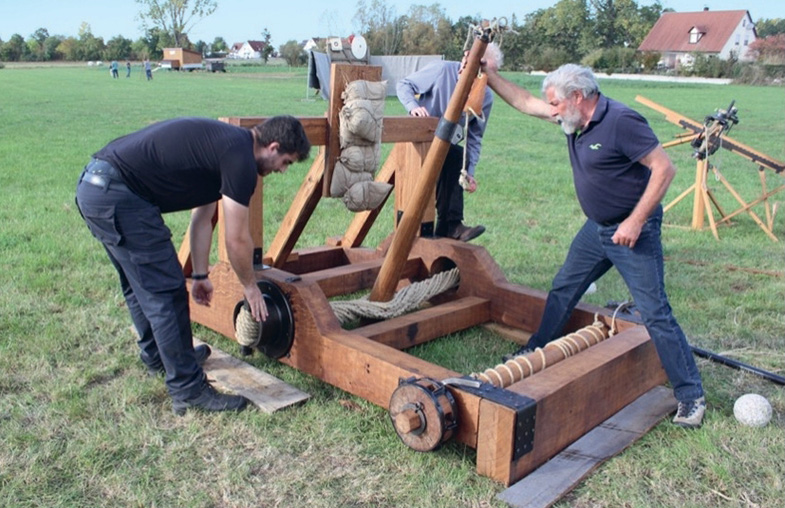 Holzkonstruktion, die von drei Männern bedient wird, auf einer flachen Wiese. Im Hintergrund Häuser und Bäume. 