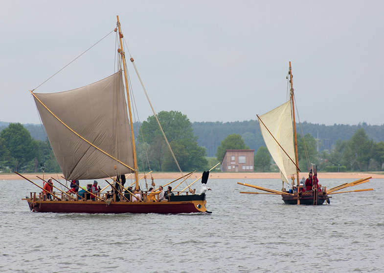 Foto zweier Segel-Ruderboote aus Holz zu Wasser, jeweils besetzt mit Menschen. Die Boote verfügen über geschnitzte Schmuckelemente (u. a. weiß-schwarzer Vogel als Gallionsfigur auf einem der beiden Boote). Im Hintergrund Ufer mit Bäumen und einem Haus.  