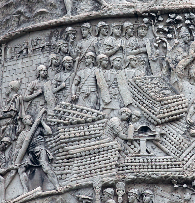 Detailaufnahme eines Wandreliefs, das römische Soldaten und Holzaufschichtungen zeigt, um welche die Soldaten versammelt sind. 