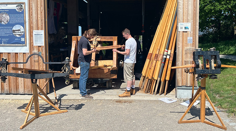 Foto einer Halle von vorne, mit geöffnetem Tor, davor zwei Geschütze, im Torbereich stehen zwei Männer vor einer weiteren Konstruktion, an die Hallenwand angelehnt stehen hohe Holzstäbe.