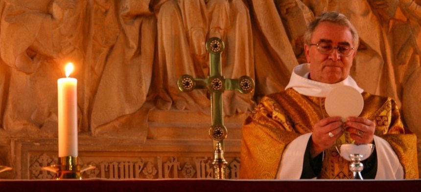 Ein Priester steht hinter dem Altar und feiert Messe. Er hält eine Hostie hoch. 