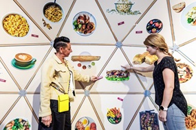 Auf einer in Dreiecke eingeteilten Ausstellungsfläche sind verschiedene Lebensmittel und Zubereitungen abgebildet, pflanzliche ebenso wie Fleisch, Gesundes und Fastfood. Davor stehen zwei Frauen und weisen auf einen Döner in der Mitte