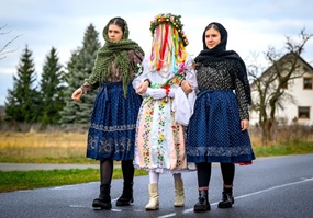 Drei junge Frauen in sorbischer Tracht, die äußeren beiden in dunklen Farben, die mittlere in weiß mit Blumen, auf ihrem Kopf ein Kranz, von dem ein dichter Schleier aus bunten Bändern herabhängt. Die beiden äußeren haben sich bei der mittleren eingehakt und führen sie über eine kleine Straße. Im Hintergrund ein Wohnhaus und Bäume.