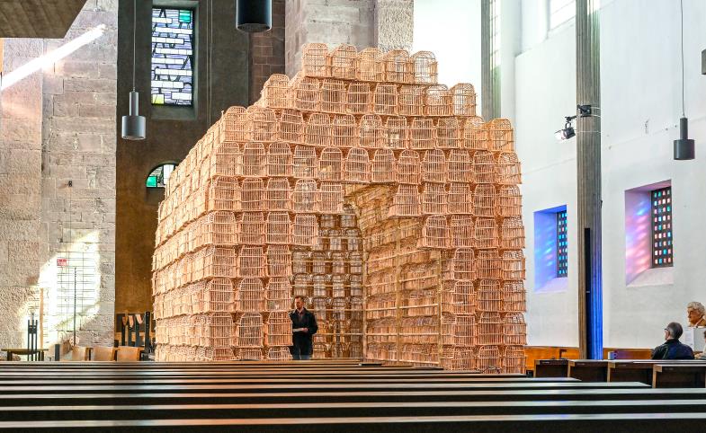 Blick in einen Kirchenraum, vor den Bänken, den Altarbereich verdeckend, eine Installation aus Vogelkäfigen, die einen Bogen formen, in den man durch ein rundes Portal vorne eintreten kann.