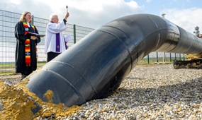 Eine schwarze Pipeline kommt aus der Erde, dahinter an einem Zaun stehen eine evangelische Pfarrerin und ein katholischer Pfarrer, letzterer mit erhobenem Aspergill