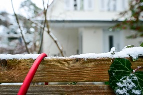 Hölzerner Gartenzaum mit Efeu im Schnee, im Hintergrund ein Einfamilienhaus. Über den Zaun läuft ein dickes rotes Kabel.