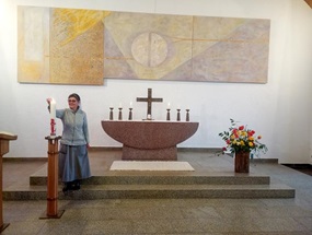 In einem modernen Kirchenraum entzündet eine Ordensschwester die Osterkerze. Im Hintergrund ein Steinaltar mit Kreuz.