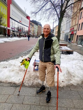 Volker Hautz, auf zwei Krücken gestützt mit sichtbaren Beinprothesen, lacht ins Bild; hinter ihm die Fußgängerzone einer Innenstadt mit Schneeresten.
