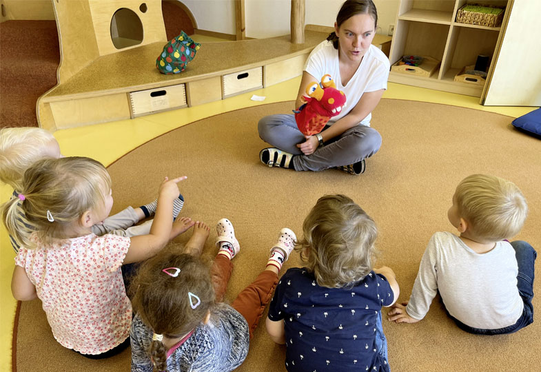 Kinder sitzen mit Erzieherin im Kreis und hören ihr zu