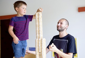 Ein Kind steht auf einem Stuhl und baut einen Turm aus Holzklötzchen, der bereits bis zu seiner Schulter reicht, während eine Fachkraft es dabei unterstützt.