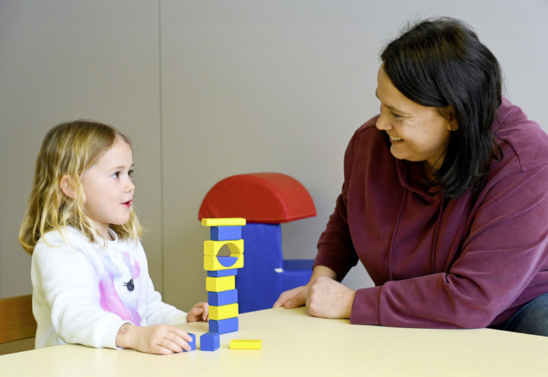 Eine Frau und ein Kind sitzen sich freundlich gegenüber, vor ihnen steht ein Turm aus blau-gelben Bauklötzchen.