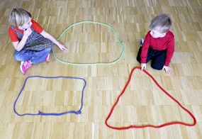 Zwei Kinder legen mit bunten Schnüren geometrische Formen auf den Boden.