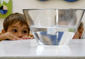 Vor einem Kind steht eine Schüssel mit Wasser und einem Eiswürfel, den das Kind aufmerksam beobachtet.