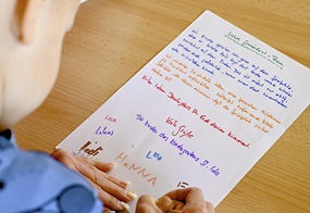 Auf einem Tisch liegt ein bunt geschriebener Brief, während ein Kind mit Unterstützung eines Erwachsenen seine Unterschrift darunter setzt.