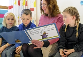 Eine Fachkraft liest drei Kindern ein Buch vor, das sie so hält, dass alle gut hineinschauen können.
