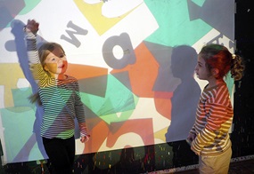 Auf einer Wand werden Buchstaben projiziert und davor stehen zwei Kinder.