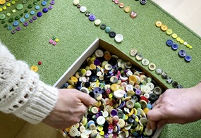 Eine Kiste mit vielen bunten Knöpfen, während zwei Hände darin suchen und einige Knöpfe nach Farben sortiert daneben liegen.