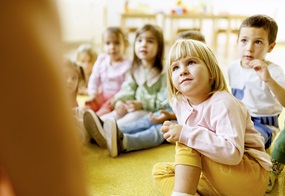 Einige Kinder sitzen auf dem Boden und blicken alle Aufmerksam nach vorne .
