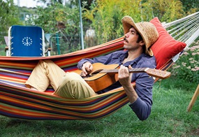 Ein Mann mit Strohhut und Gitarre sitzt in einer bunten Hängematte.