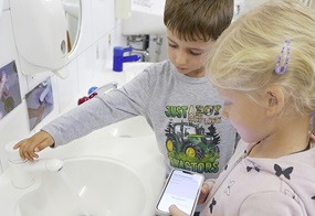 Ein Kind öffnet den Wasserhahn, während das andere Kind mit dem Handy auf aufnehmen drückt. 