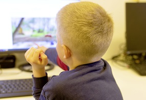 Ein Kind ist von hinten zu sehen und zeigt mit der rechten Hand auf den Bildschirm eines Computers.