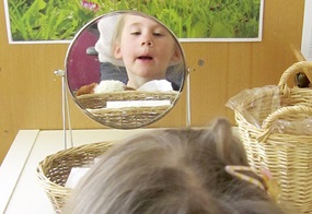 Auf einem Tisch stehen ein Spiegel und Taschentücher, und ein Kind blickt in den Spiegel, kurz bevor es sich die Nase putzt.
